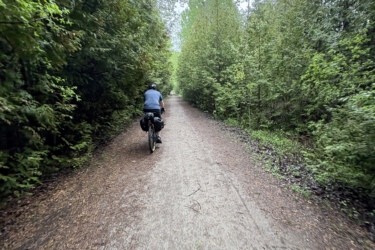 A cyclist riding on a gravel path surrounded by lush greenery on both sides, with trees lining the trail and a cloudy sky overhead. The perspective captures the view from slightly behind the cyclist, emphasizing the peaceful, natural setting. The Great Trail: Uxbridge mountain bike trail.
