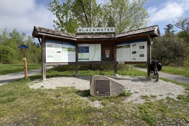 Information kiosk for Blackwater Area, featuring various panels with details about the location, surrounded by greenery and a walking path. A stone with a plaque is visible in front, with a bicycle parked nearby. The Great Trail: Uxbridge mountain bike trail.