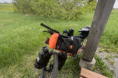 A mountain bike with gear attached is resting against a wooden shelter. The bike has an orange and black bag and water bottle, set against a backdrop of green grass and a bush under a partly cloudy sky. A wooden bench is visible in the foreground.