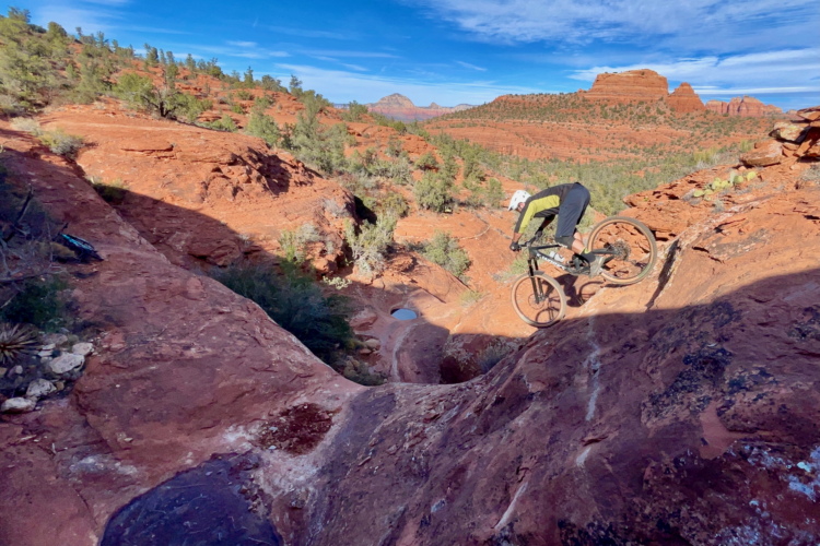 A mountain biker navigates a rocky trail in a desert landscape, capturing air as they perform a jump. The scene features red rock formations and vegetation under a clear blue sky.