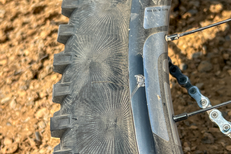 Close-up of a mountain bike tire showing tread patterns and dirt accumulation, with part of the bike frame and chain visible in the background. The ground appears rocky and textured.