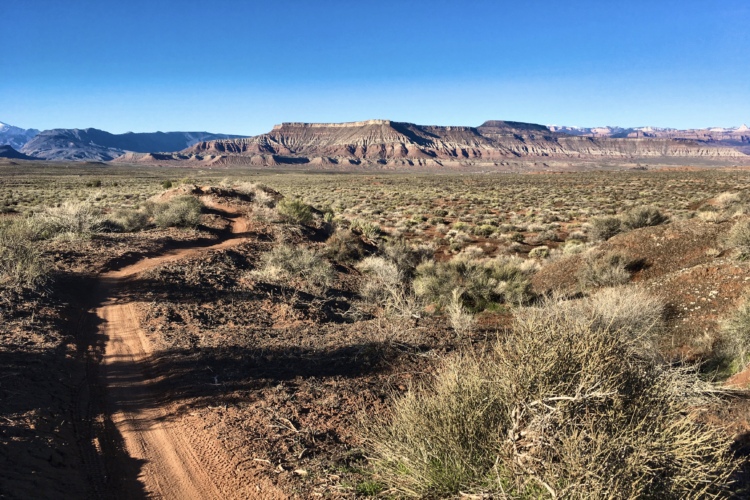 A scenic view of a dirt path winding through a rugged landscape, with sparse vegetation and a backdrop of layered cliffs and mountains under a clear blue sky.