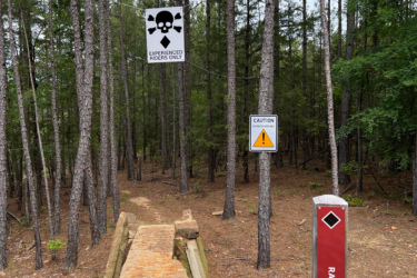 A narrow dirt path leads into a wooded area, marked by a "Caution" sign and a large sign reading "EXPERIENCED RIDERS ONLY" featuring a skull and crossbones. To the right, a trail marker sign is labeled "RAVEN." The landscape is filled with tall pine trees and a mix of dirt and rocks lining the trail. Red Lick Trails mountain bike trail.