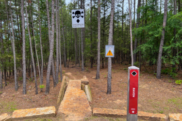 A narrow dirt path leads into a wooded area, flanked by tall pine trees. Two signs are visible: one warns "EXPERIENCED RIDERS ONLY" with a skull and crossbones symbol, and the other displays a caution symbol. A marker post labeled "RAVEN" stands to the right, indicating the trail name. The ground is covered with fallen pine needles and scattered rocks. Red Lick Trails mountain bike trail.