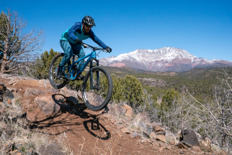 A mountain biker airborne over a rocky trail, with a backdrop of snow-capped mountains and a clear blue sky. The cyclist is wearing a helmet and athletic gear in shades of blue, demonstrating a dynamic jump on the bike. Surrounding greenery and rugged terrain create a natural setting for outdoor adventure.