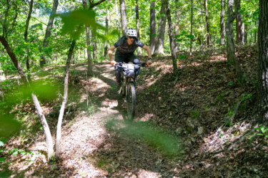 A mountain biker rides on a narrow dirt trail surrounded by lush greenery and trees, demonstrating skill and control as they navigate the terrain. Sunlight filters through the leaves, creating a vibrant outdoor atmosphere. WOKA Whitewater Park mountain bike trail.