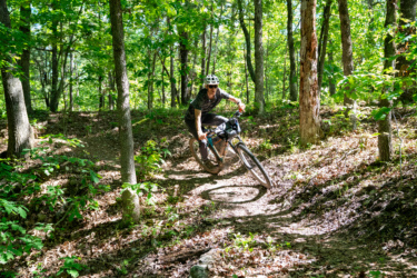 A mountain biker navigating a winding trail in a lush green forest, leaning into a turn with trees and foliage surrounding the path. The cyclist is wearing a helmet and riding a bike equipped with a front suspension. Sunlight filters through the leaves, creating a dappled light effect on the ground. WOKA Whitewater Park mountain bike trail.