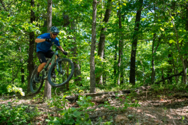 A mountain biker performing a jump on a forest trail surrounded by lush green trees and undergrowth. The cyclist is wearing a helmet and is airborne above a small log on the trail. WOKA Whitewater Park mountain bike trail.
