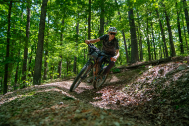 A mountain biker navigating a winding trail in a lush green forest, with trees surrounding the path. The rider is wearing a helmet and sunglasses and is leaning into the curve while pedaling on a sunny day. WOKA Whitewater Park mountain bike trail.