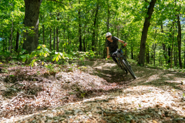 A cyclist wearing a helmet and sunglasses rides a mountain bike along a winding trail in a lush green forest, surrounded by tall trees and dappled sunlight filtering through the leaves. The ground is covered with a mix of dirt, leaves, and small rocks. WOKA Whitewater Park mountain bike trail.