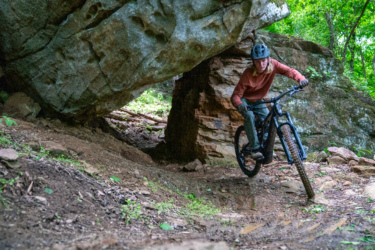 A person riding a mountain bike through a narrow passage under a large rock formation in a forested area. The rider, wearing a helmet and casual clothing, appears focused as they navigate the trail surrounded by trees and natural terrain. Horseshoe Canyon Ranch mountain bike trail.