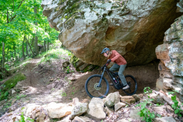 A mountain biker navigates a rocky trail beneath a large boulder in a lush forest. The cyclist, wearing a helmet and casual outdoor attire, is focused on maintaining balance on their bike as they maneuver through the rugged terrain surrounded by greenery. Horseshoe Canyon Ranch mountain bike trail.