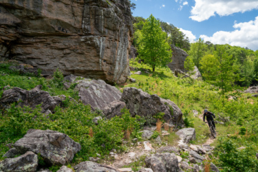 A mountain biker navigates a rocky trail amidst lush greenery and towering cliffs under a partly cloudy sky. Horseshoe Canyon Ranch mountain bike trail.