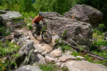 A person riding an electric mountain bike on a rocky trail surrounded by greenery and large boulders. The rider, wearing a helmet and gloves, navigates the terrain with caution under a clear blue sky. Horseshoe Canyon Ranch mountain bike trail.
