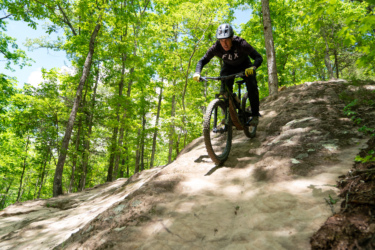 A mountain biker navigating a rocky downhill trail in a lush green forest, wearing a helmet and gloves, with sunlight filtering through the trees. The biker is focused on maintaining balance as they ride over the uneven terrain. Horseshoe Canyon Ranch mountain bike trail.