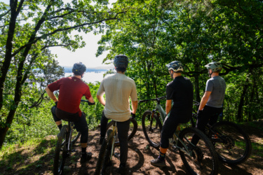 Four mountain bikers are standing on a wooded trail, facing a scenic view of a body of water. The cyclists are wearing helmets and varying styles of cycling attire, with their bikes parked beside them. The surrounding area is lush with greenery, and the sunlight filters through the trees, creating a serene outdoor atmosphere. Gravity Falls mountain bike trail.