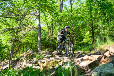 A mountain biker navigating a rocky trail in a lush, green forest during a sunny day. The cyclist is focused and wearing a helmet and protective gear. Trees and greenery surround the pathway, highlighting an outdoor adventure setting. River Mountain mountain bike trail.