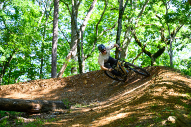 A mountain biker skillfully navigates a dirt trail through a lush green forest, leaning into a turn on a well-maintained berm. Sunlight filters through the trees, illuminating the rider and the path. Gravity Falls mountain bike trail.