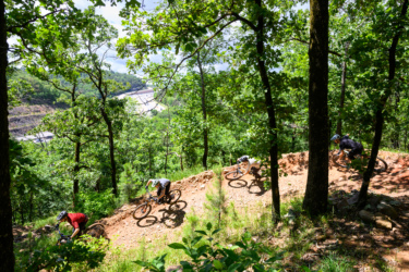 A group of four mountain bikers navigating a dirt trail through a dense, green forest. The trail winds downward, and trees provide a lush backdrop. Sunlight filters through the leaves, highlighting the texture of the path and the riders' movements. In the distance, a glimpse of a road can be seen, adding context to the outdoor adventure. River Mountain mountain bike trail.
