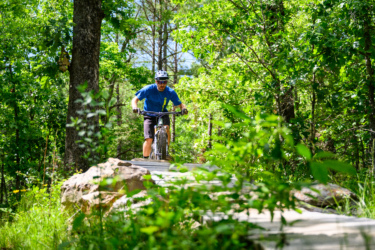 A mountain biker navigating a wooden trail surrounded by lush greenery in a forested area. The rider is wearing a blue shirt, helmet, and sunglasses, focused on the path ahead while riding on a narrow boardwalk. Dhu Drop mountain bike trail.