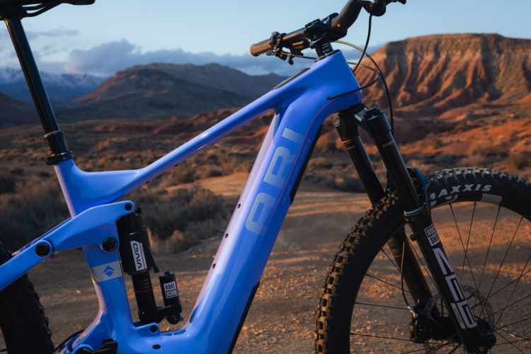 A close-up of a blue mountain bike frame against a natural landscape, featuring rugged hills and a sunset sky. The bike highlights include the suspension system, handlebars, and tires, showcasing its design and functionality. The background presents a scenic view of varying terrain.