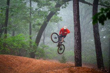 A mountain biker in a bright orange jacket performs a jump over a dirt mound in a misty forest, surrounded by tall trees and greenery. Chaotic Zone mountain bike trail.