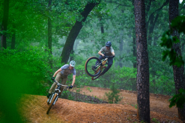 Two mountain bikers performing tricks on a dirt trail in a forested area. One rider is jumping off a ramp, while the other leans into a turn, surrounded by lush green trees and foliage. The scene captures a dynamic moment in a natural setting with overcast lighting. Chaotic Zone mountain bike trail.