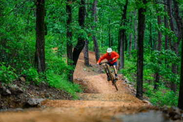A mountain biker in an orange jacket and white helmet performs a jump on a dirt trail, surrounded by lush green trees and foliage. The trail winds through a forest, showcasing a vibrant outdoor setting. Pinnacle Mountain State Park mountain bike trail.