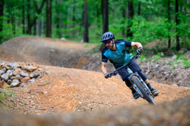 A mountain biker wearing a helmet and protective gear is leaning into a turn on a dirt trail in a wooded area. The trail features mounds of dirt and rocks, surrounded by lush green trees. Chaotic Zone mountain bike trail.
