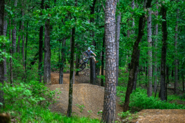 A mountain biker performs a mid-air trick over a dirt jump, surrounded by dense green trees in a forested area. Chaotic Zone mountain bike trail.