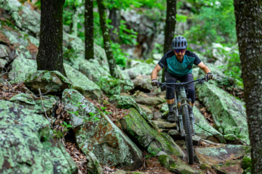 A mountain biker navigating a rocky trail surrounded by trees in a lush green forest. The rider, wearing a helmet and sunglasses, is focused on maintaining balance while maneuvering over the uneven terrain. Pinnacle Mountain State Park mountain bike trail.