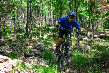 A mountain biker riding a trail through a forest, navigating rocky terrain with greenery surrounding him. The cyclist is wearing a blue shirt, black shorts, a helmet, and sunglasses, showcasing an active outdoor lifestyle. Dhu Drop mountain bike trail.