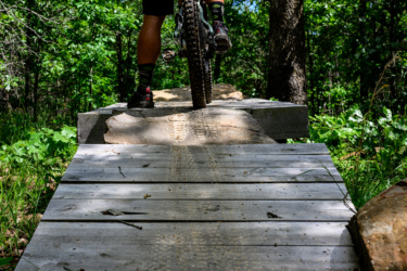 A close-up view of a mountain biker's foot on a wooden bike ramp surrounded by lush green vegetation and trees in a forested area. The ramp features natural elements, with portions made from wood and stone, highlighting the outdoor biking trail. Dhu Drop mountain bike trail.