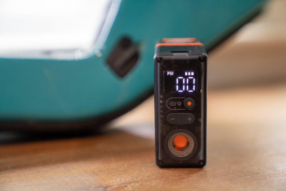 Digital air pressure gauge displaying a reading of 0 PSI, positioned on a wooden surface with a blurred teal object in the background. The gauge features an LED screen and several control buttons.
