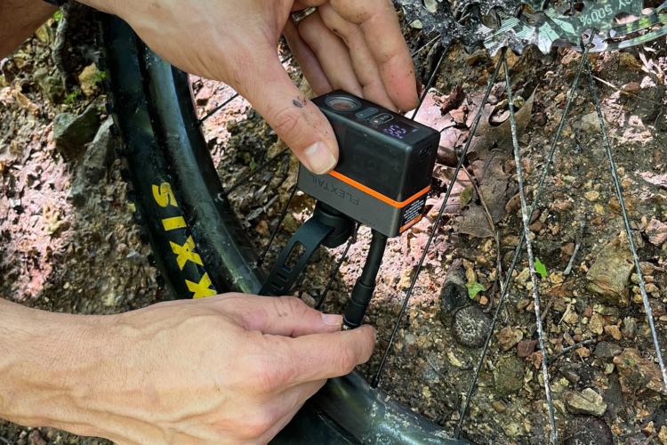 A close-up of a person's hands using a digital tire pressure gauge to check the pressure of a bicycle tire. The bike's tire is resting on a gravelly surface, and the gauge is connected to the valve stem. The tire features a yellow logo, and dirt is visible on the hands, indicating an outdoor cycling environment.