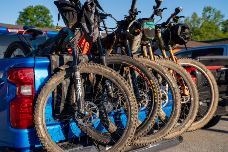 A close-up view of several mountain bikes secured in the back of a blue pickup truck, showing their muddy tires and gear bags. The bikes are lined up, showcasing various colors and styles against a backdrop of trees and vehicles.
