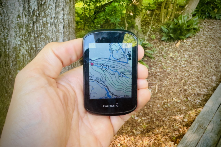 A hand holding a Garmin GPS device displaying a detailed map of a hiking trail, surrounded by greenery in a forested area.