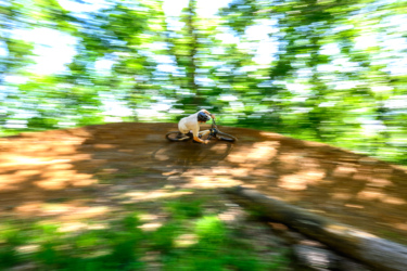 A mountain biker expertly navigates a dirt track, leaning into a turn with a blur of motion and greenery surrounding them. Sunlight filters through the trees, creating a vibrant backdrop for the action. Gravity Falls mountain bike trail.