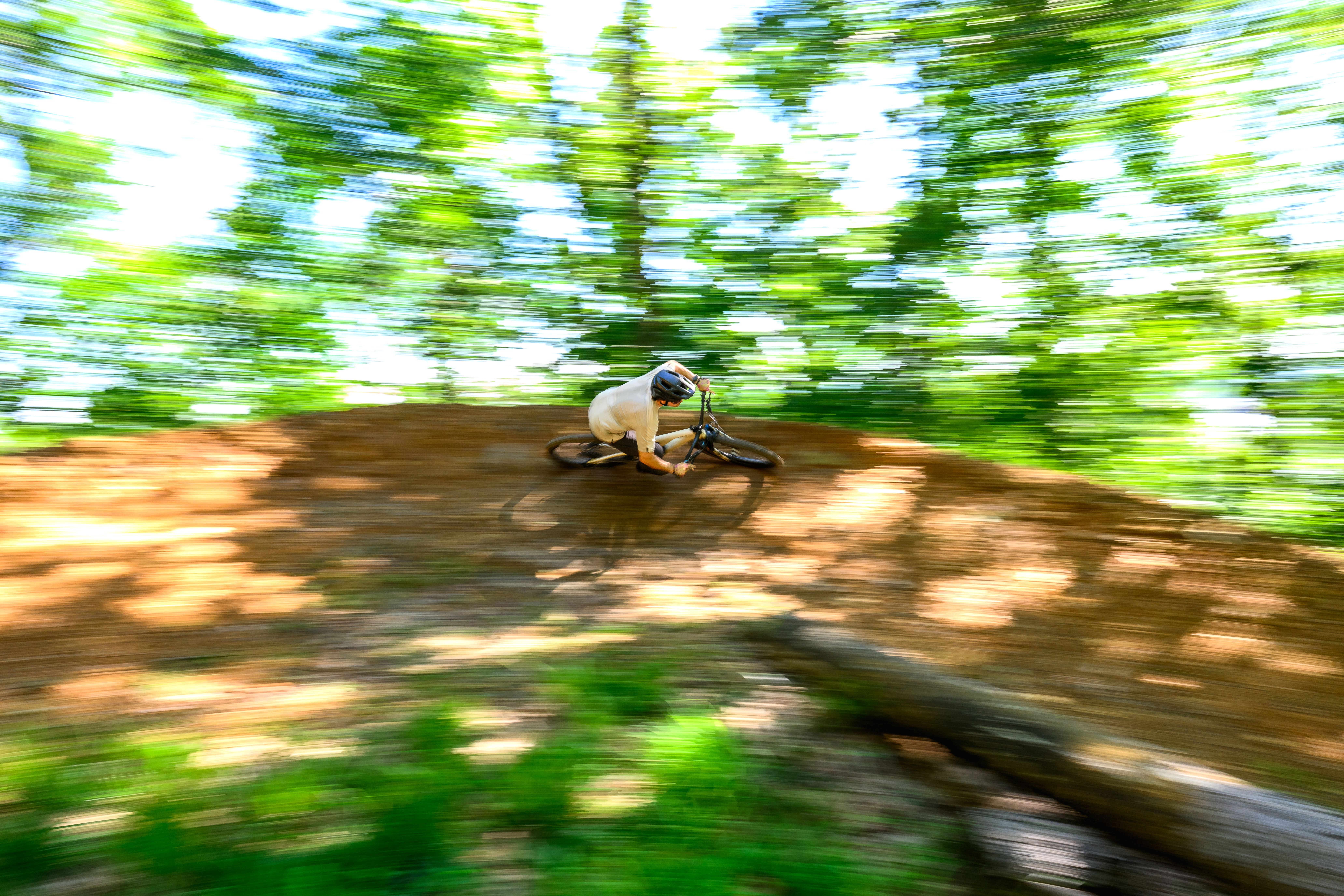 A mountain biker expertly navigates a dirt track, leaning into a turn with a blur of motion and greenery surrounding them. Sunlight filters through the trees, creating a vibrant backdrop for the action. Gravity Falls mountain bike trail.