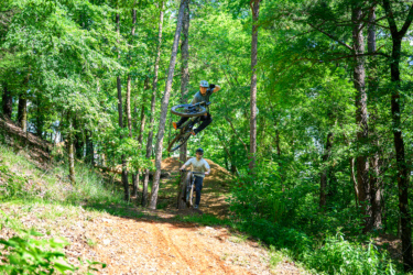 Two mountain bikers in a lush green forest setting. One rider is performing a jump over a dirt mound, capturing the air with their bike, while the other rider is positioned on the ground, observing. The scene showcases a vibrant natural environment with trees and foliage. River Mountain mountain bike trail.