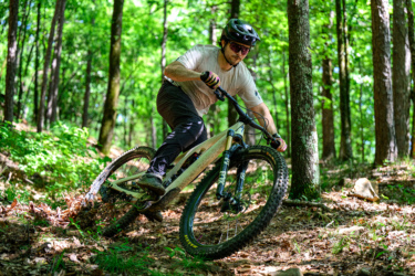 A mountain biker dressed in a light shirt and dark pants navigates a curved trail in a vibrant green forest, leaning into the turn on a light-colored mountain bike. The ground is covered with fallen leaves and surrounded by trees, creating a dynamic outdoor scene. River Mountain mountain bike trail.