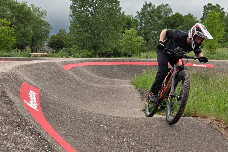 A cyclist in a black shirt and helmet rides a mountain bike on a pump track, navigating a curved section of the path marked with a red stripe. Lush greenery surrounds the track under a cloudy sky.