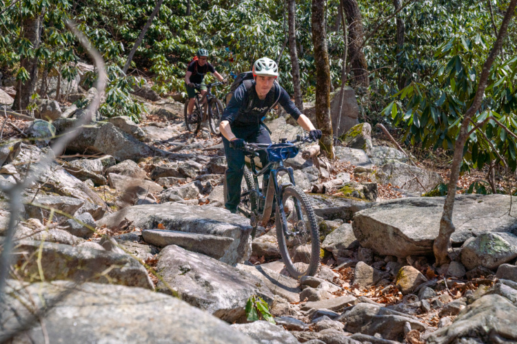 Two mountain bikers navigate a rocky trail through a wooded area. The foreground features one cyclist focused on maneuvering over large stones, while a second rider follows in the background. The scene is set among lush greenery and trees, indicating a vibrant outdoor environment.