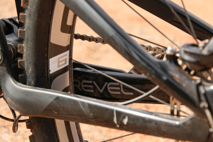 Close-up view of a mountain bike's drivetrain and frame, showing the chain, gear system, and branding on the bike frame, with a blurred background of earthy terrain.