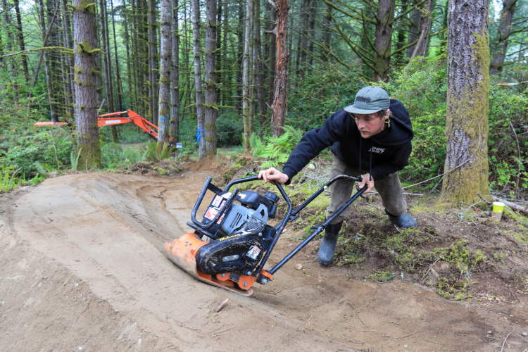 A man operating a gas-powered compactor on a dirt path in a wooded area, surrounded by tall trees and shrubs.