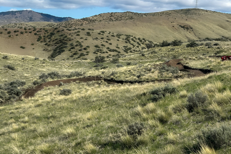 A winding dirt path cuts through a grassy landscape with rolling hills and sparse vegetation, under a mostly cloudy sky. In the background, a power line intersects the horizon, while distant mountains rise in the background.