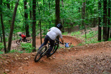 A mountain biker navigating a winding dirt trail in a lush green forest, with another cyclist visible in the background. The rider is leaning into a turn, showcasing skillful handling of the bike, while the scene is filled with trees and foliage. River Mountain mountain bike trail.