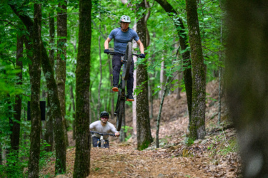 Two mountain bikers ride through a forested trail. One rider is performing a jump with both wheels off the ground, while the other follows behind on a bike. The scene is set against a backdrop of green trees and earthy terrain. River Mountain mountain bike trail.