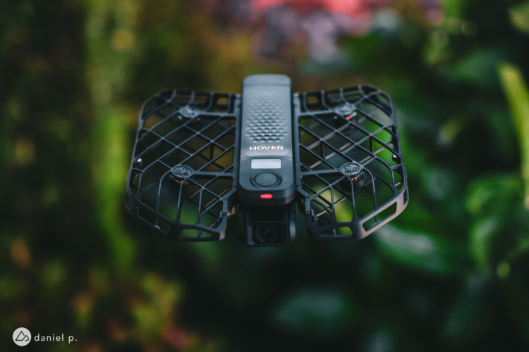 A black drone with a protective cage hovering in a green, blurred outdoor setting. The drone features a compact design with a camera and control buttons visible on its surface.