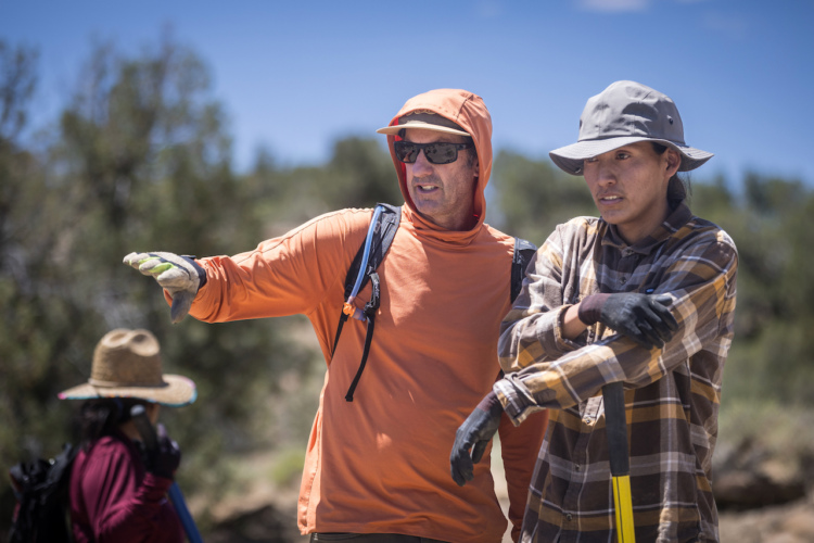 Two individuals engaged in a discussion outdoors, with one gesturing while wearing an orange long-sleeve shirt and a hood. The other person, dressed in a checkered shirt and a gray hat, appears to be listening thoughtfully. In the background, a person wearing a straw hat is partially visible. The setting is sunny with a natural landscape.